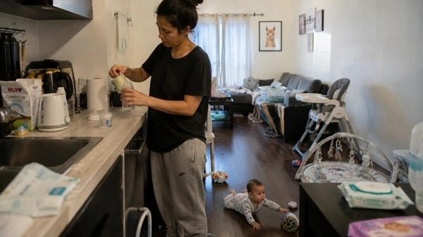 Sleepy parent trying to prepare a bottle while a 6-month-old baby reaches toward objects on the floor in a cluttered kitchen-living area