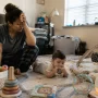 Parent sitting on a messy living room floor with a 6-month-old baby holding a teether and scattered baby toys around them in natural morning light
