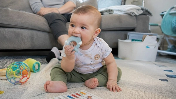 6-month-old baby chewing a silicone teether while sitting on a play mat with a parent nearby watching casually