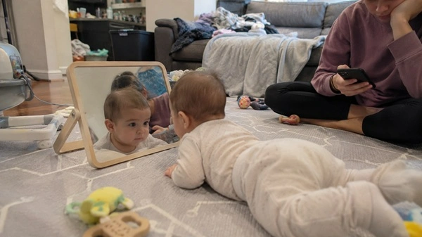 Baby doing tummy time on a play mat facing a floor mirror while parent sits nearby scrolling on phone